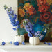 Two white vases with blue flowers on a white surface in front of a colorful floral painting.