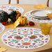 A round white placemats with a colorful autumn harvest pattern featuring pumpkins, acorns, leaves, and floral motifs, styled on a wooden dining table with plates, a glass, and fall decorations.