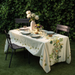 A rectangular table laid with a natural-colored tablecloth featuring floral patterns, with dishes and a vase of flowers on top, set against a lush greenery backdrop.