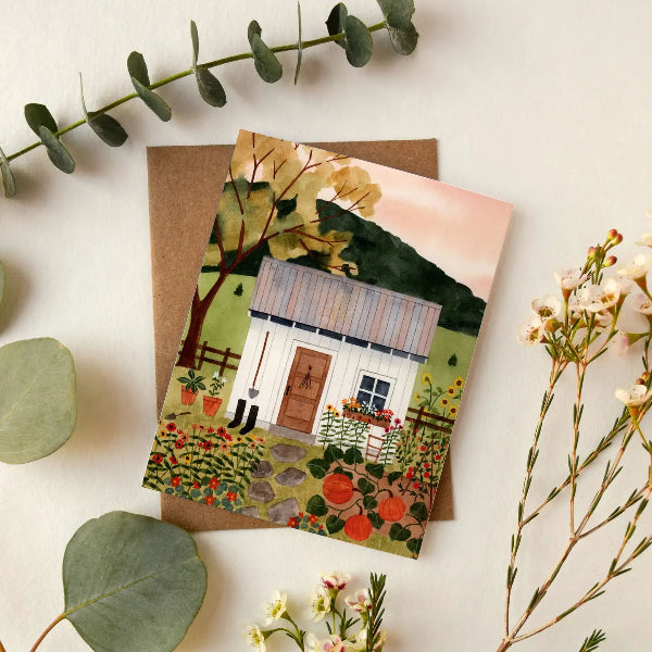 A square art print or greeting card featuring a painted country cabin surrounded by flowers and trees, styled on a kraft envelope with eucalyptus leaves and small flowers on a light neutral background.