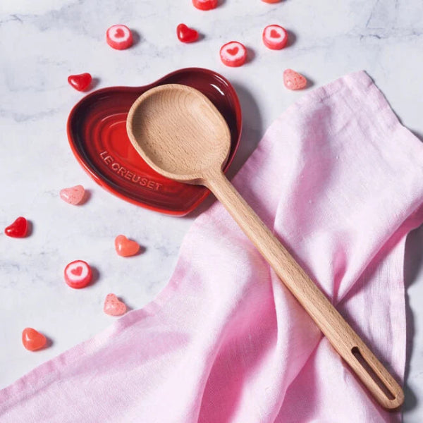 Wooden spoon on a pink cloth with heart-shaped candies and a Le Creuset heart-shaped dish in the background.