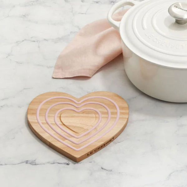Heart-shaped wooden trivet with concentric circles on a marble surface next to a white pot.