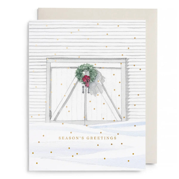 A holiday greeting card with a watercolor-style white barn door, green wreath, red bow, and scattered gold dots, shown with a beige envelope behind it on a white background.