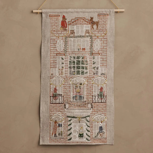 A vertical woven wall hanging tapestry featuring a detailed multi-story building facade with windows, figures, and decorative motifs, suspended from a light wood rod against a plain beige wall.