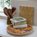A styled kitchen scene with a stack of cookbooks on a decorative round tray, next to a utensil holder and a textured wooden board leaning against a white marble wall.