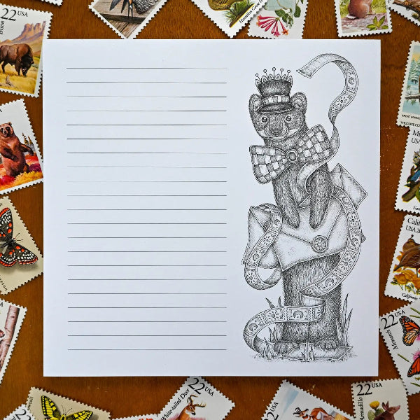 A lined paper notepad featuring a black-and-white sketch of a bear wearing a top hat and bow tie, surrounded by colorful postage stamps on a wooden surface.