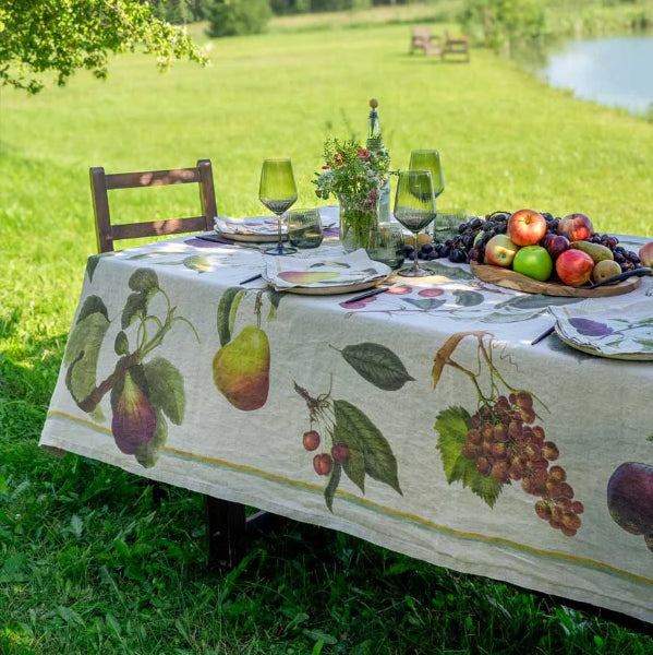 A rustic outdoor dining table set in a grassy field near a lake, featuring a linen tablecloth printed with detailed illustrations of pears, grapes, cherries, and plums. The table is styled with plates, green wine glasses, a small floral centerpiece, and a wooden fruit basket filled with fresh fruit.