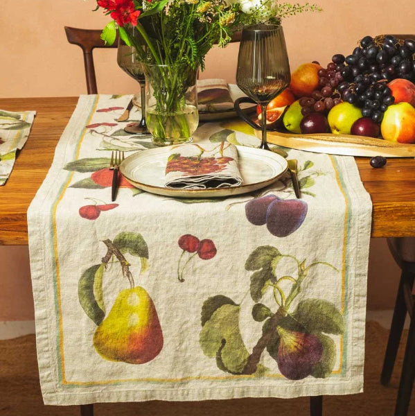 A rectangular linen table runner with a vintage fruit print, styled on a wooden dining table with a floral centerpiece, wine glass, and fruit platter against a warm peach background.