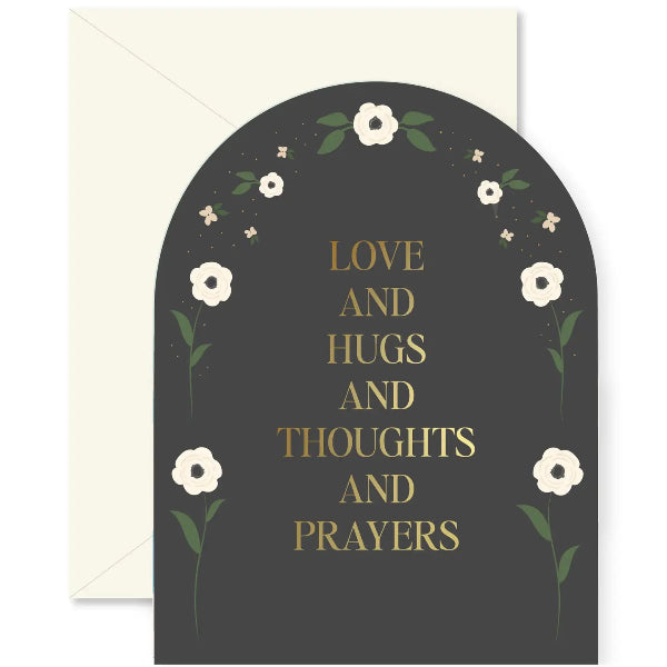 A greeting card with a dark charcoal arched background, featuring the words 'LOVE AND HUGS AND THOUGHTS AND PRAYERS' in gold lettering, surrounded by delicate white flowers and green leaves. A cream-colored envelope is partially visible behind the card.
