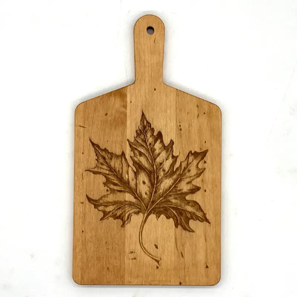 A rectangular wooden cutting board with a handle, featuring a laser-burned maple leaf design in the center, shown against a plain white background.