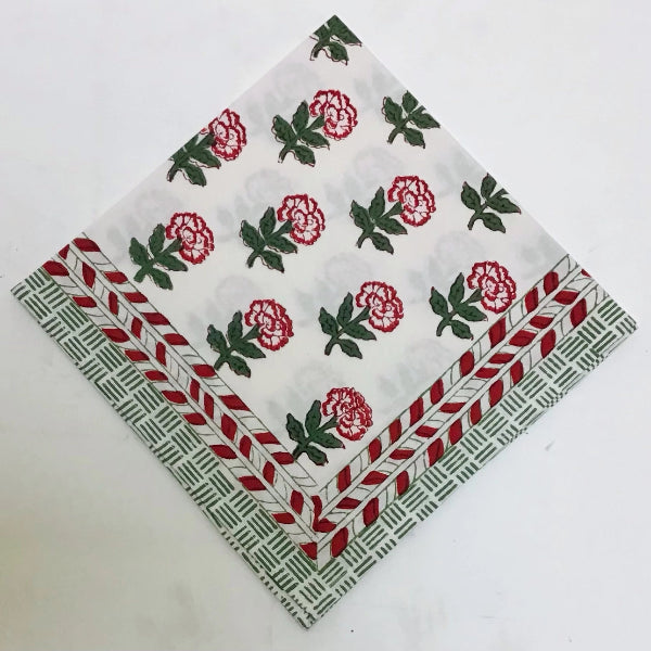 A square white fabric napkin featuring a red and green floral block print and a decorative border, laid flat on a white surface.