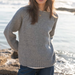 A woman wearing a light gray knit sweater and blue jeans, standing outdoors near the water with sunlight reflecting on the sea.