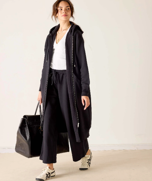 A woman models a long black zip-up hooded jacket over a white top and black wide-leg pants, holding a black tote bag against a plain off-white studio background.