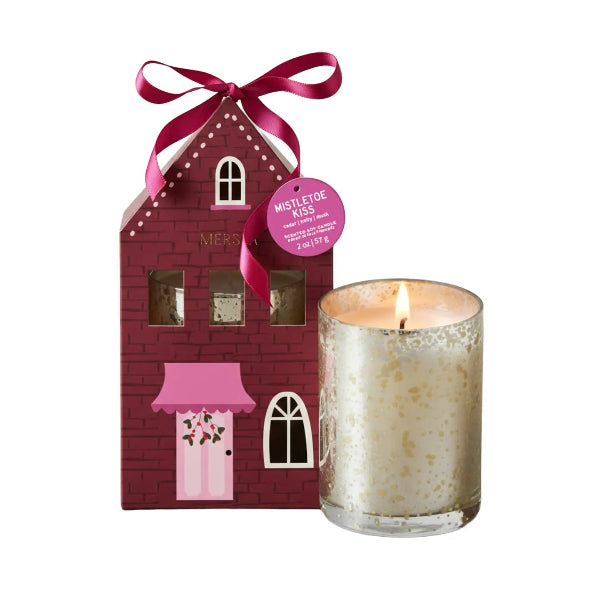A holiday-themed scented candle set featuring a 'Mistletoe Kiss' candle in a clear glass jar with gold glitter and a lit flame, next to its red brick house-shaped packaging box with a pink bow and purple circular tag.
