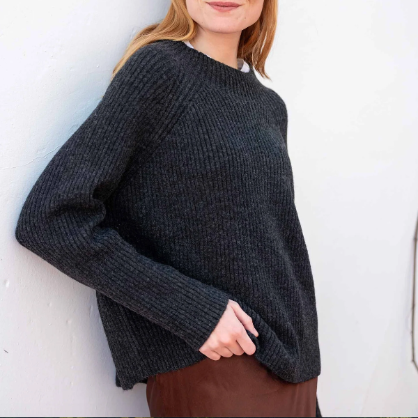 A woman wearing a dark charcoal gray ribbed knit sweater, leaning against a white wall.