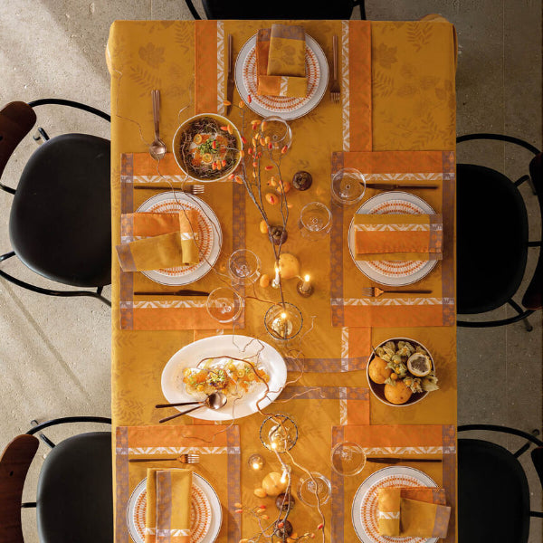 An overhead view of a dining table set with a warm orange and yellow tablecloth, white plates with orange patterns, folded napkins, wine glasses, and autumn-themed centerpieces, surrounded by black chairs.