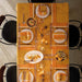 An overhead view of a dining table set with a warm orange and yellow tablecloth, white plates with orange patterns, folded napkins, wine glasses, and autumn-themed centerpieces, surrounded by black chairs.