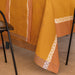 A mustard yellow tablecloth with geometric and paisley border patterns draped over a table, shown in a real-world setting with the legs of black chairs visible.