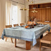 A light blue floral tablecloth with a blue patterned border is draped over a wooden dining table set with chairs, candles, and decorative items in a warm, sunlit dining room.
