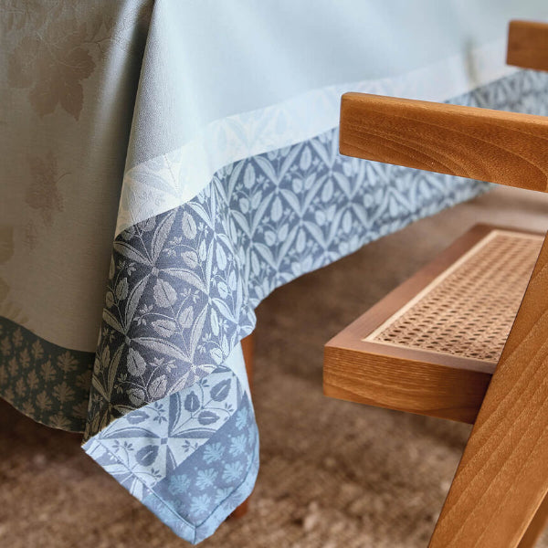 A close-up of a patterned tablecloth draped over a table, showing a light blue and gray botanical print with a blue-gray border, next to a wooden chair with a cane seat.