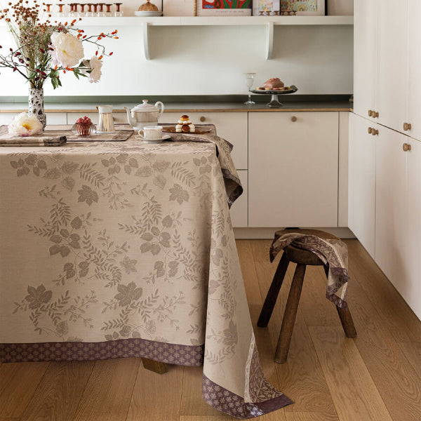 A beige floral tablecloth with a brown leaf pattern and a patterned border is draped over a dining table in a bright, modern kitchen setting.