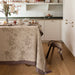 A beige floral tablecloth with a brown leaf pattern and a patterned border is draped over a dining table in a bright, modern kitchen setting.