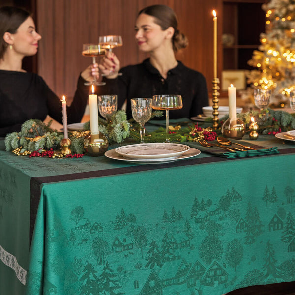 A festive holiday table setting with a teal green tablecloth printed with a winter village pattern, decorated with candles, plates, and pine garlands, with two women holding wine glasses in the background.
