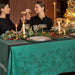 A festive holiday table setting with a teal green tablecloth printed with a winter village pattern, decorated with candles, plates, and pine garlands, with two women holding wine glasses in the background.
