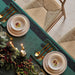 An overhead view of a styled holiday table setting with a deep teal table runner, matching napkins, gold flatware, white ceramic bowls, and a festive evergreen garland with red berries.