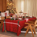 A festive dining table set for a holiday gathering, featuring a red tablecloth with green and white patterned borders, matching red napkins, white plates, and lit taper candles. The scene is decorated with a Christmas tree, gift boxes, and wooden chairs in a warmly lit indoor setting.