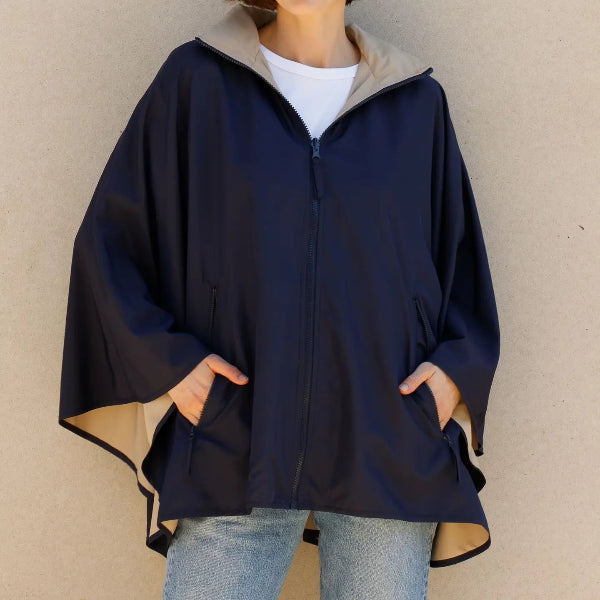 A woman wearing a navy blue hooded poncho-style jacket with a beige inner lining, layered over a white t-shirt and light blue jeans, standing against a neutral beige wall.