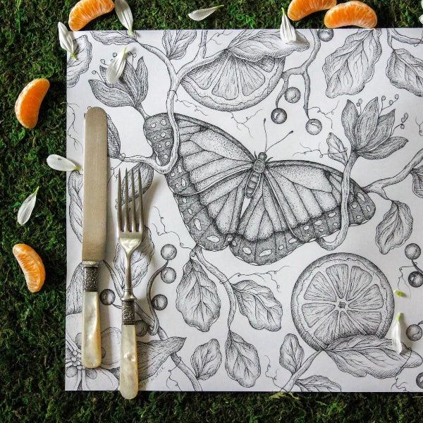 A square white placemat with a detailed black and white botanical illustration featuring a large butterfly, leaves, berries, and citrus slices, styled on a green grass-like surface with a knife, fork, and small orange slices.