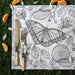 A square white placemat with a detailed black and white botanical illustration featuring a large butterfly, leaves, berries, and citrus slices, styled on a green grass-like surface with a knife, fork, and small orange slices.