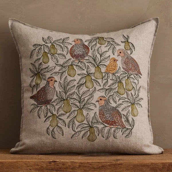 A square linen throw pillow with a detailed embroidered design of birds and pears, resting on a rustic wooden surface against a textured beige wall.