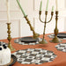 Halloween-themed table setting with patterned placemats, antique-style brass candelabras, white taper candles, and a white cake on a rust-colored tablecloth.