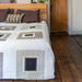 A bed dressed in a cream and taupe patchwork quilt with geometric square patterns, styled with brown and striped pillows in a bedroom with a light wood headboard, white walls, and dark wood flooring.
