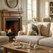 A cozy living room scene with a beige upholstered sofa featuring a decorative throw pillow with a bird and leaf design, set in front of a lit fireplace. A wooden coffee table with teacups and pastries sits in the foreground, and a matching armchair is partially visible on the left.