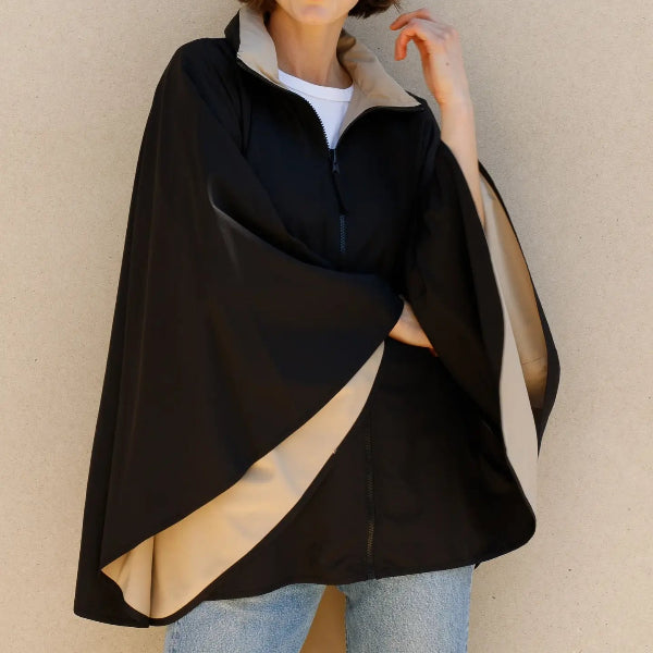 A woman wearing a black cape-style zip-up jacket with a beige inner lining and collar, paired with light blue jeans, standing against a neutral beige wall.