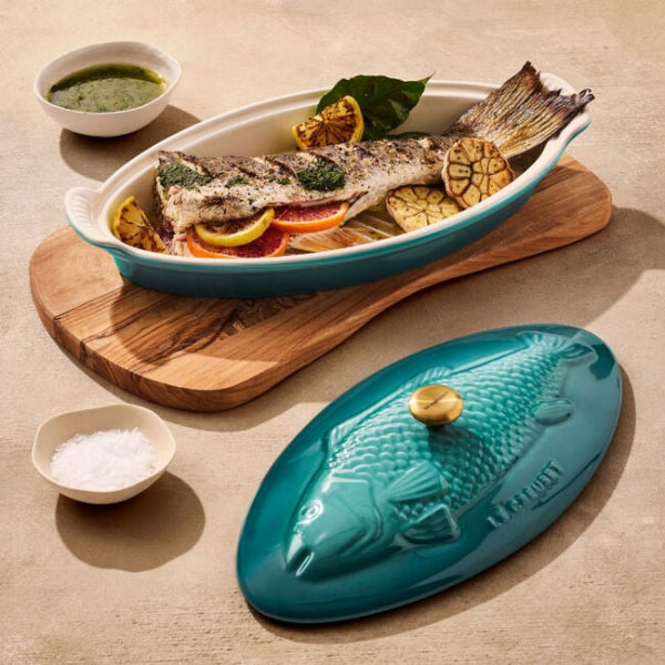Teal fish-shaped ceramic dish with cooked fish and vegetables on a wooden board.