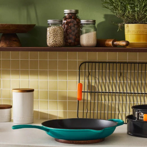 Teal frying pan on a kitchen counter with jars and a shelf in the background.