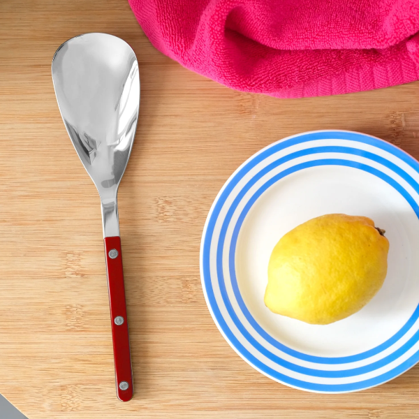 A stainless steel serving spoon with a reddish-brown wooden handle lies on a light wood surface next to a white plate with blue stripes holding a yellow lemon, with a pink cloth in the top corner.