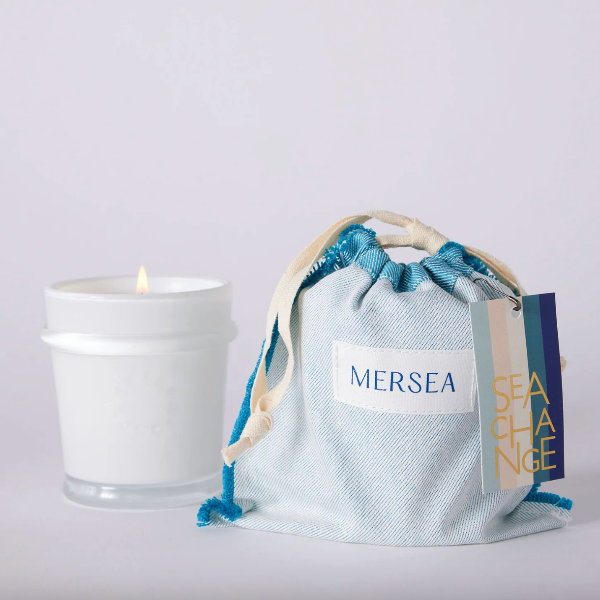 White candle next to a blue drawstring bag with 'MERSEA' branding on a light gray background
