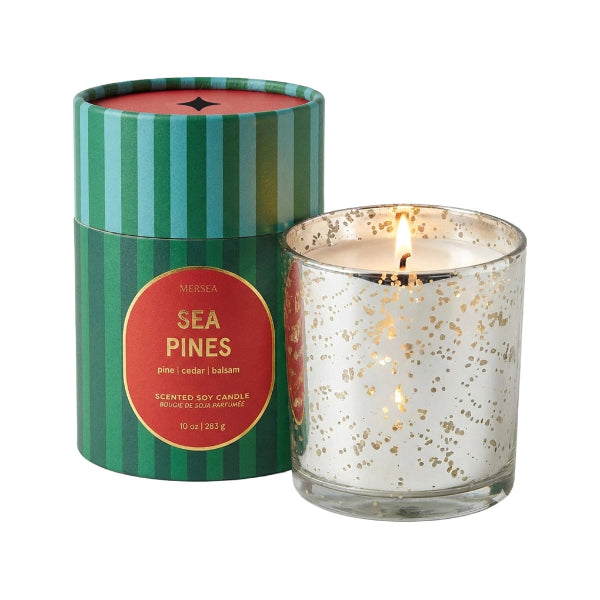 A lit scented soy candle in a clear glass jar with gold speckles sits next to its green and teal striped cylindrical packaging box labeled 'Mercea Sea Pines' on a white background.