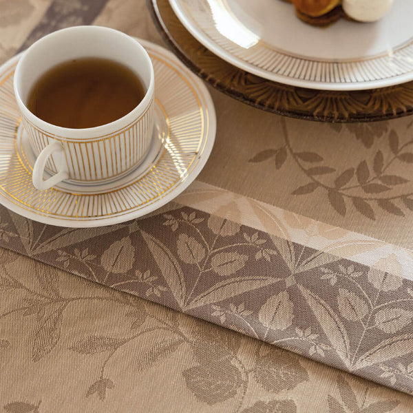 A close-up of a beige and taupe floral jacquard table runner with a matching folded napkin, styled with a white teacup of tea on a gold-rimmed saucer and plates.