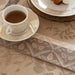 A close-up of a beige and taupe floral jacquard table runner with a matching folded napkin, styled with a white teacup of tea on a gold-rimmed saucer and plates.