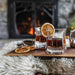 Two glasses of whiskey with orange slices on a wooden tray in front of a fireplace.