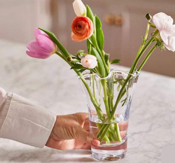 Person holding a clear glass vase with colorful flowers on a light background