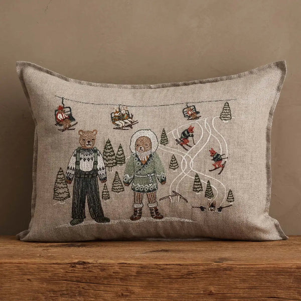 A square linen throw pillow with a rustic embroidered ski resort scene, resting on a wooden surface against a neutral beige wall.