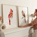 Woman arranging two framed artworks of colorful birds on a white wall.