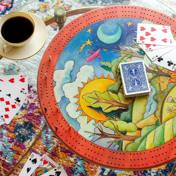 A round wooden board game table with a colorful illustrated scene on top, surrounded by playing cards and a cup of black coffee on a patterned rug.
